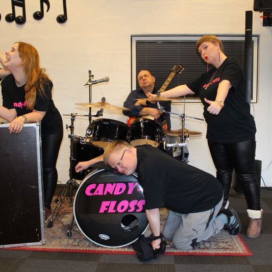 Fem personer står ved et trommesæt. Der står "Candy-floss" på trommesættet, og personerne har matchende trøjer.