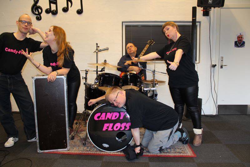 Fem personer står ved et trommesæt. Der står "Candy-floss" på trommesættet, og personerne har matchende trøjer.