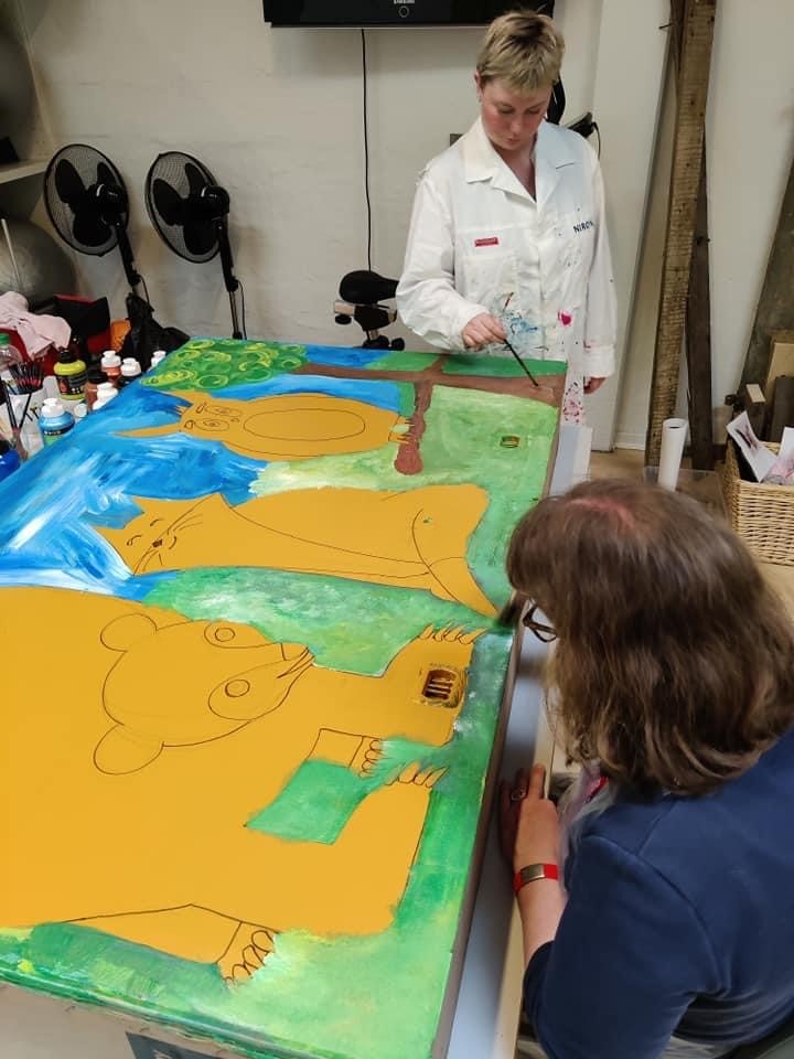 Maleri af tre forskellige dyr med en baggrund af græs og blå himmel. Det er en bjørn, kat og ugle.