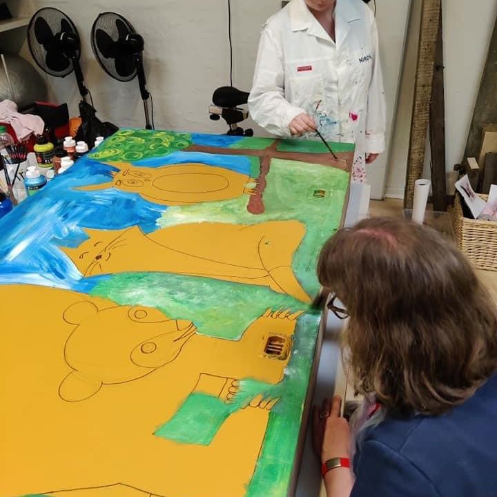 Maleri af tre forskellige dyr med en baggrund af græs og blå himmel. Det er en bjørn, kat og ugle.